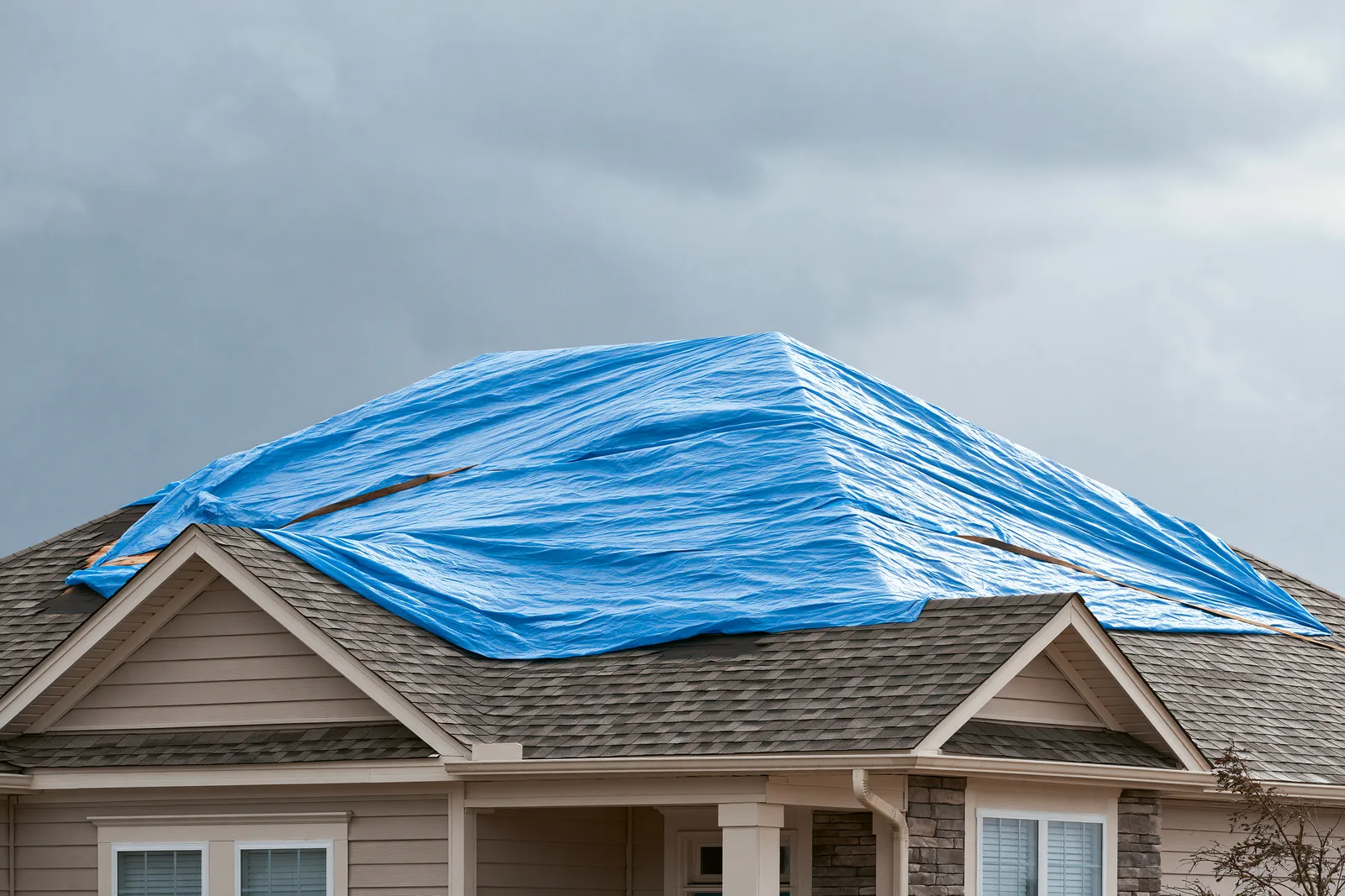 A blue tarp covers storm-damaged roof sections on a two-story home under grey clouds